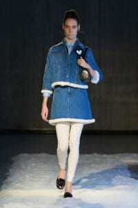 Model walking a snowy runway in a blue denim coat with white fur trim, floral-patterned white leggings, and black sandals while carrying a black bag.