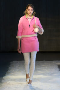 Model walking a snowy runway in a pink coat with white fur trim, white patterned tights, and white heeled shoes, carrying a round pink bag and pearl necklace