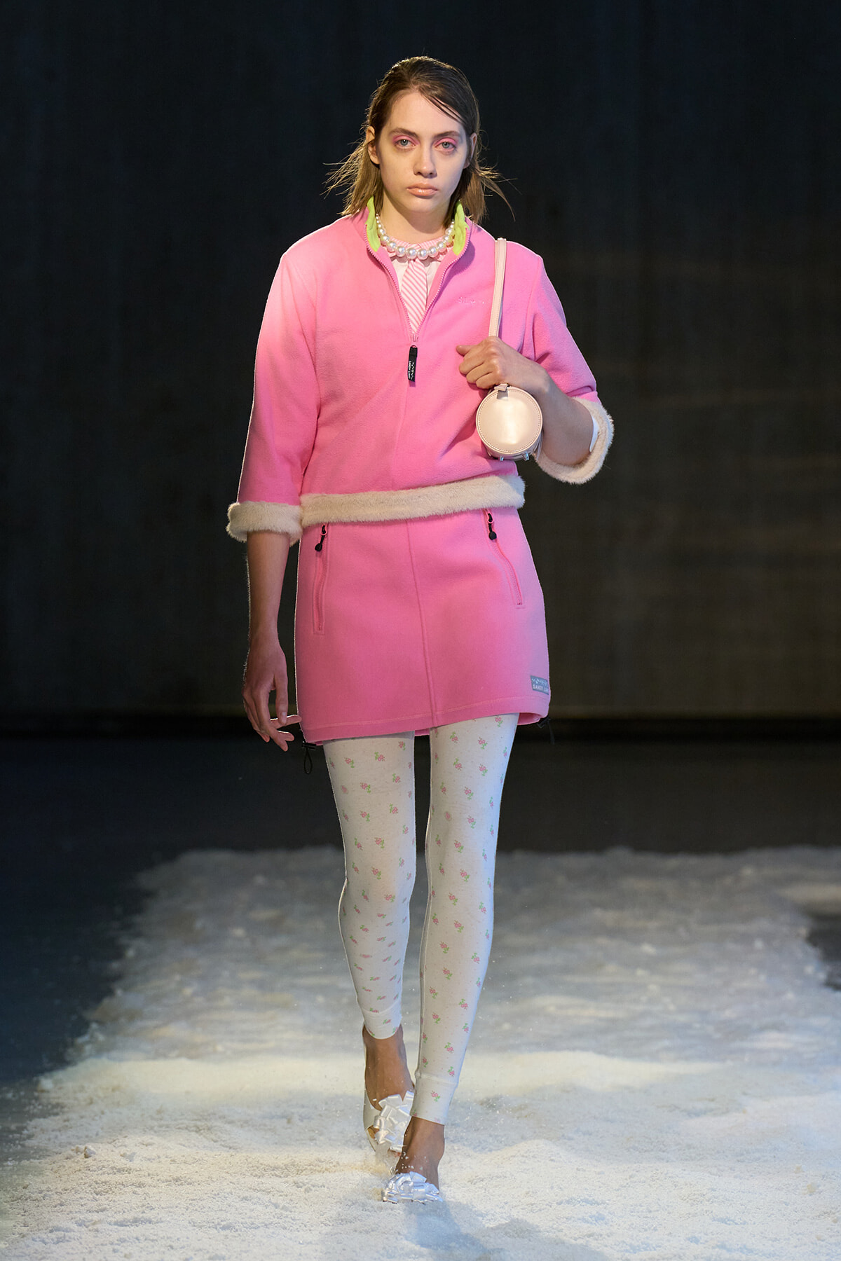 Model walking a snowy runway in a pink coat with white fur trim, white patterned tights, and white heeled shoes, carrying a round pink bag and pearl necklace