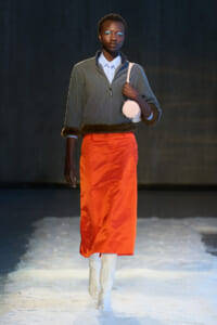Model walking a fashion runway in a gray zip sweater with fur trim, bright orange skirt, white boots, and a round pink bag over the shoulder on a snowy floor.