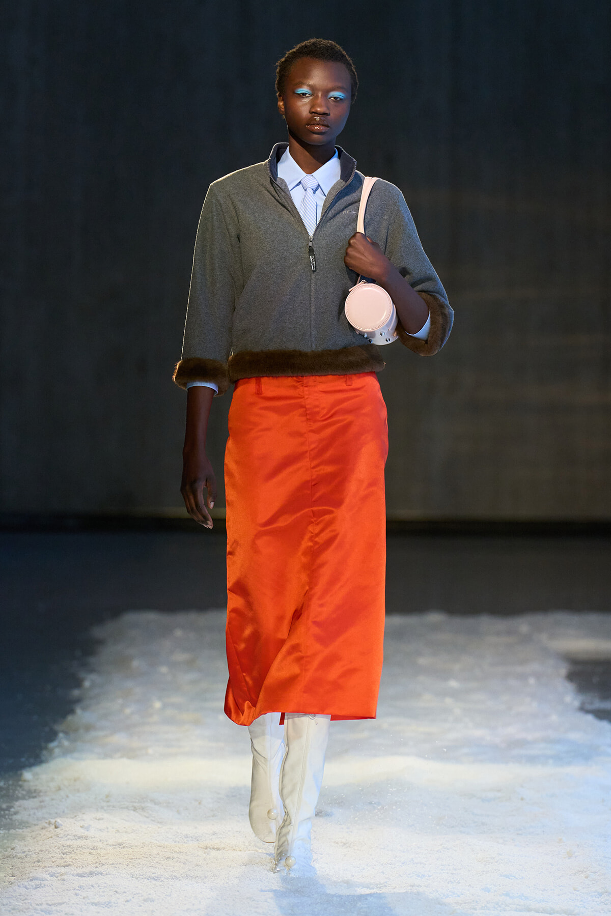 Model walking a fashion runway in a gray zip sweater with fur trim, bright orange skirt, white boots, and a round pink bag over the shoulder on a snowy floor.