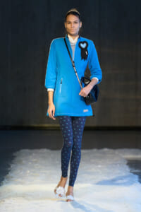 Model walking a runway in a vivid blue coat with a black heart badge, navy patterned leggings, white floral sandals, and a black crossbody bag.