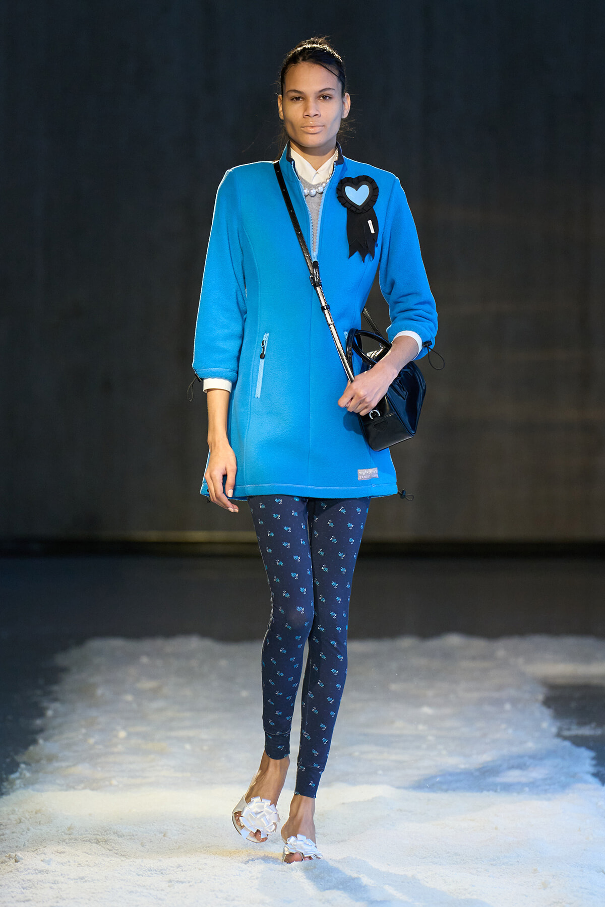 Model walking a runway in a vivid blue coat with a black heart badge, navy patterned leggings, white floral sandals, and a black crossbody bag.