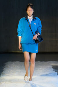 Model walking a fashion show in a bright blue oversized jacket and matching skirt, carrying a black handbag and wearing a pearl necklace.