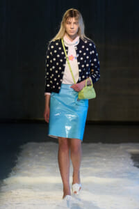 Model walking runway in navy polka-dot cardigan, pink shirt, and light blue patent skirt, holding lime handbag