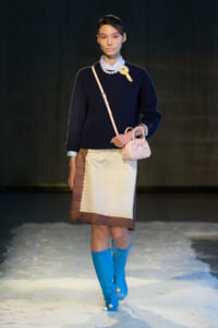 Model on a snowy runway wearing a navy sweater, cream apron, pink crossbody bag, and bright blue knee-high boots.