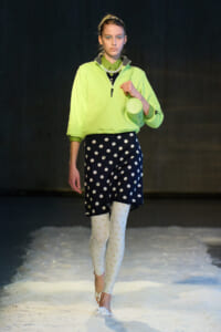 Model walking the runway in a neon lime pullover with a navy polka-dot skirt, white floral tights, and white heeled sandals, carrying a small lime bag.