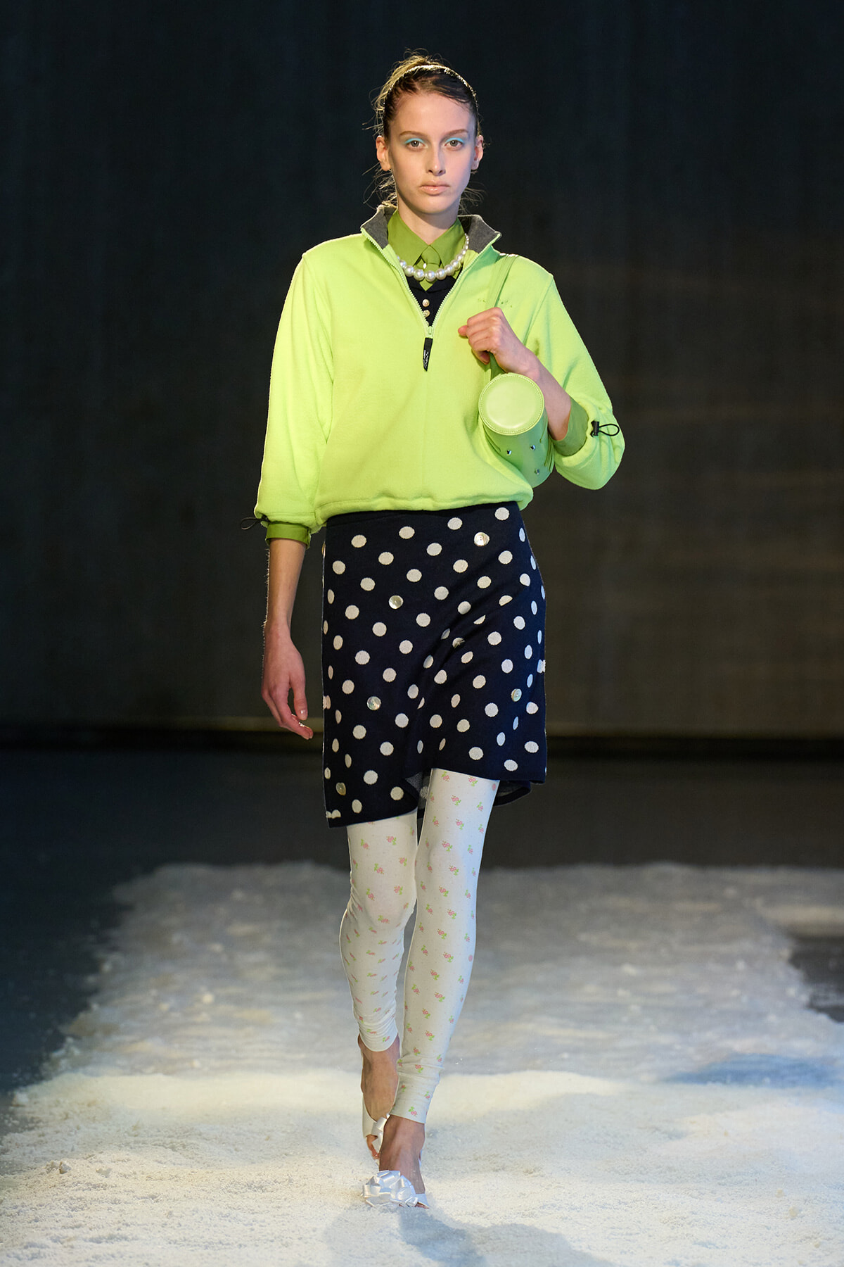 Model walking the runway in a neon lime pullover with a navy polka-dot skirt, white floral tights, and white heeled sandals, carrying a small lime bag.