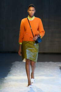 Model on a runway wearing an orange knit sweater, lime collar, glossy olive skirt, pearl necklace, and a small blue crossbody bag.