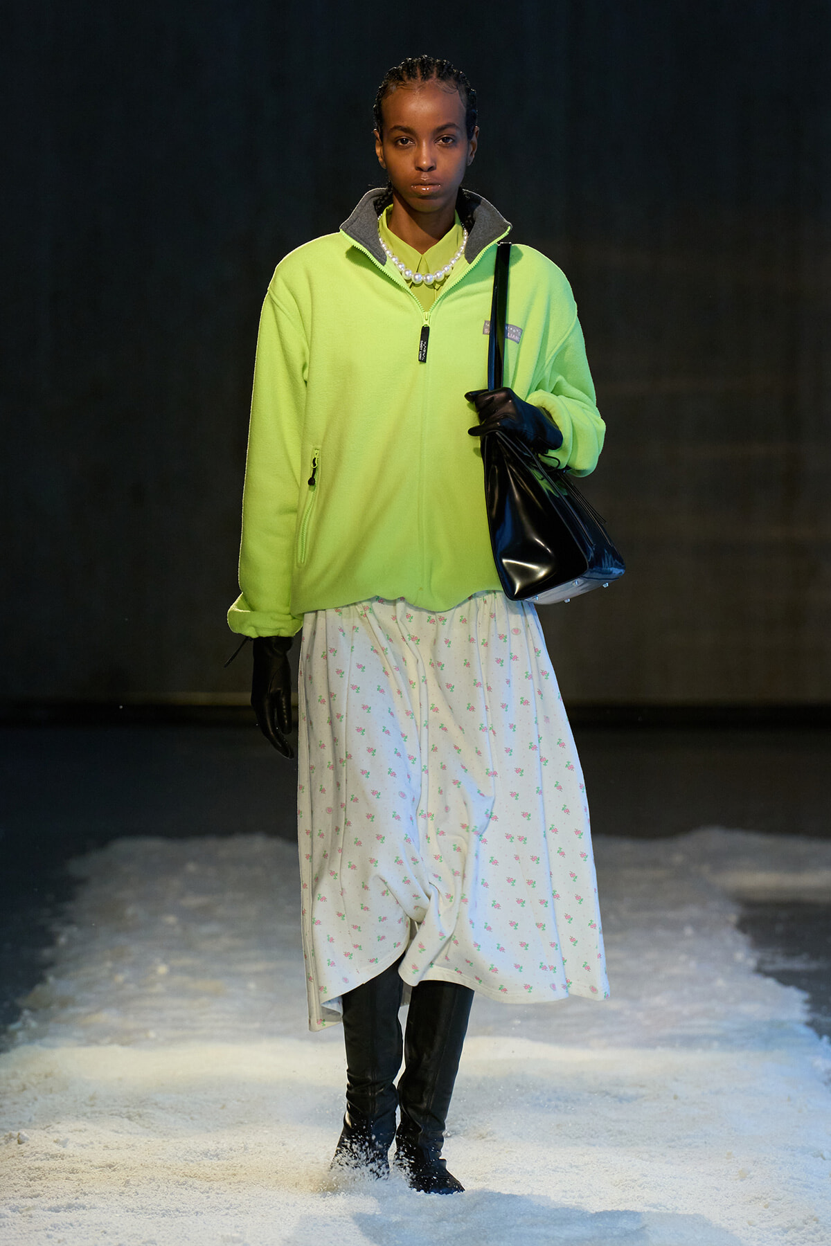 Model on a snowy runway wearing a neon lime fleece jacket, white floral skirt, black gloves, and a black handbag.
