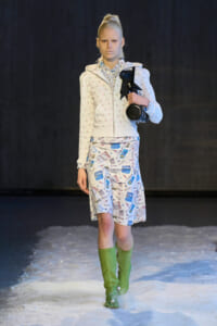 Model walking a fashion runway in a white patterned hoodie, matching print skirt, and bright green knee-high boots on a snowy runway, carrying a round black bag.