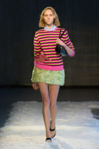 Fashion model walking a runway in a bright pink, black, and yellow striped sweater with a mint-green fuzzy mini skirt, carrying a black round handbag.