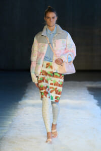 Model walking a runway in a pastel quilted jacket, graphic-print skirt, light tights, and a pale pink handbag.