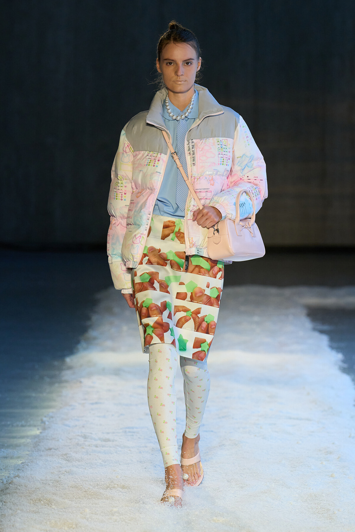 Model walking a runway in a pastel quilted jacket, graphic-print skirt, light tights, and a pale pink handbag.