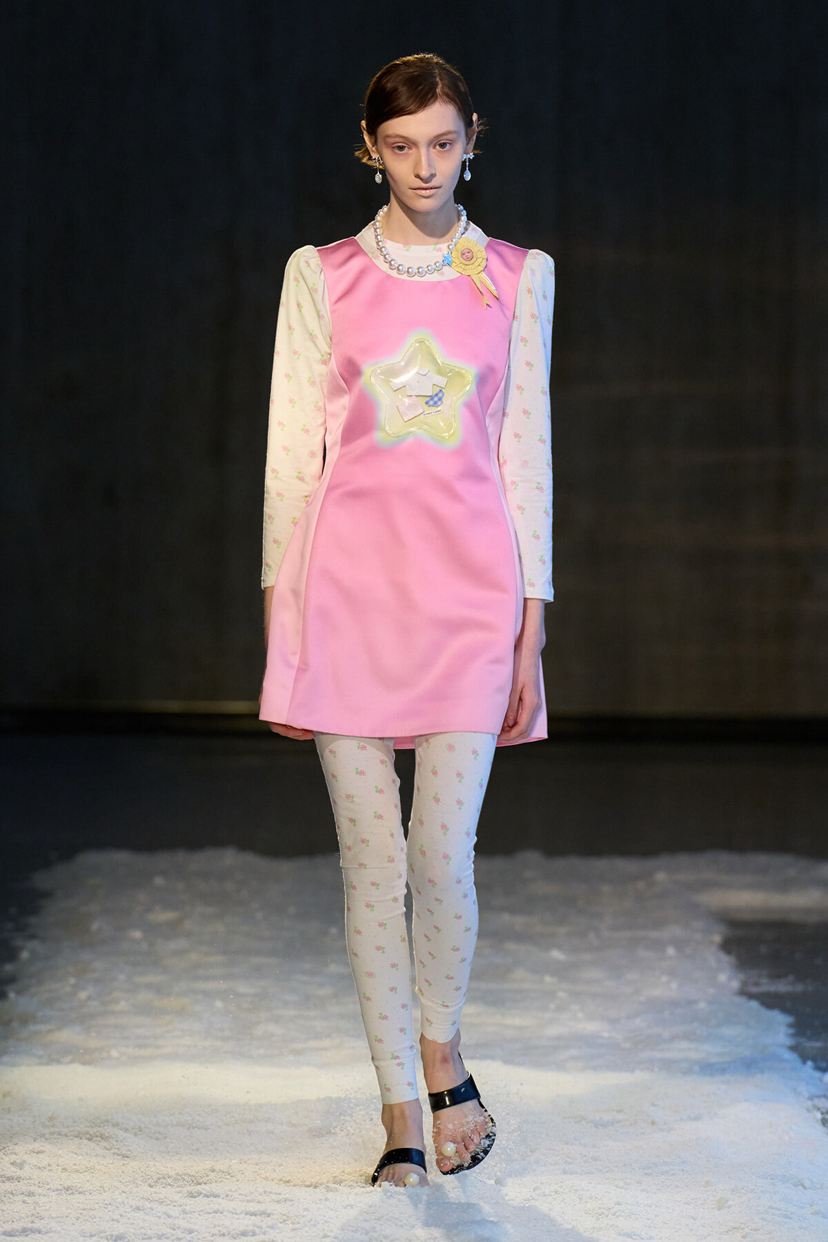 Model wearing a pink shift dress with star pocket, pearl necklace, and floral long sleeves on a snowy runway.'