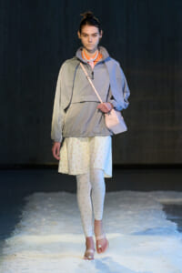 Model walking a runway in a light gray windbreaker, pale pink crossbody bag, white floral leggings, and pale pink sandals on a snowy runway, with a pearl necklace.