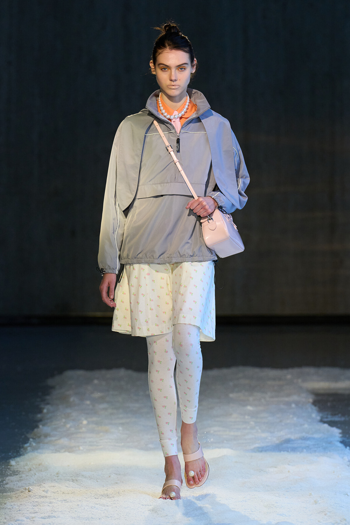 Model walking a runway in a light gray windbreaker, pale pink crossbody bag, white floral leggings, and pale pink sandals on a snowy runway, with a pearl necklace.