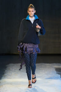 Model walks a runway wearing a navy fleece zip-up with blue collar, a ruffled polka-dot skirt, patterned leggings, and black sandals with white beads.