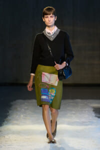 Runway model in a black sweater with a pink shirt, pearl necklace, olive skirt printed with laundry-item graphics, and a small black handbag.