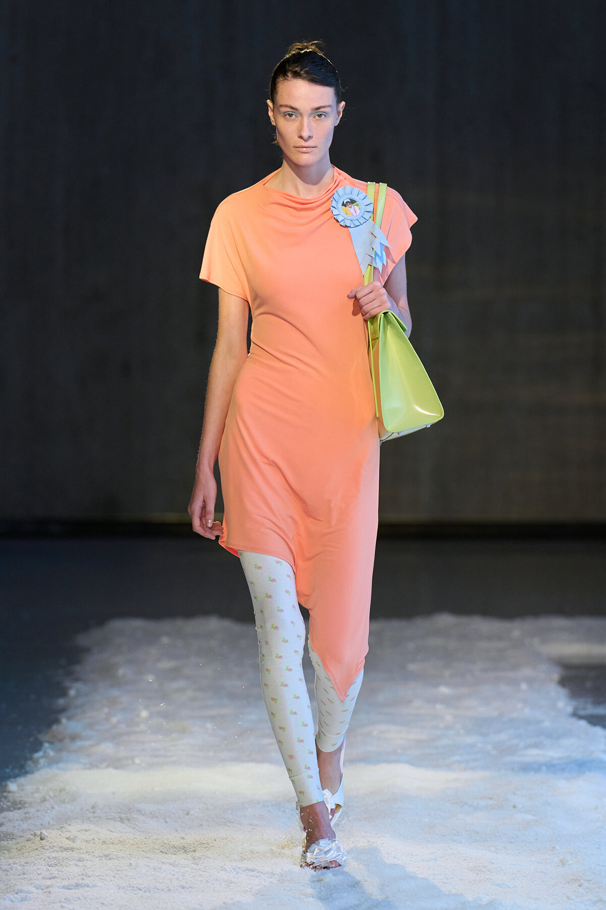 Model walks a peach midi dress with light patterned white tights and white sandals, carrying a lime tote bag with a blue ribbon badge badge on shoulder.