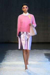 Model walking a runway in a pink knit sweater, pearl necklace, ribbon badge, iridescent pink-metallic skirt, white shoes, and light pink bag with sunglasses.