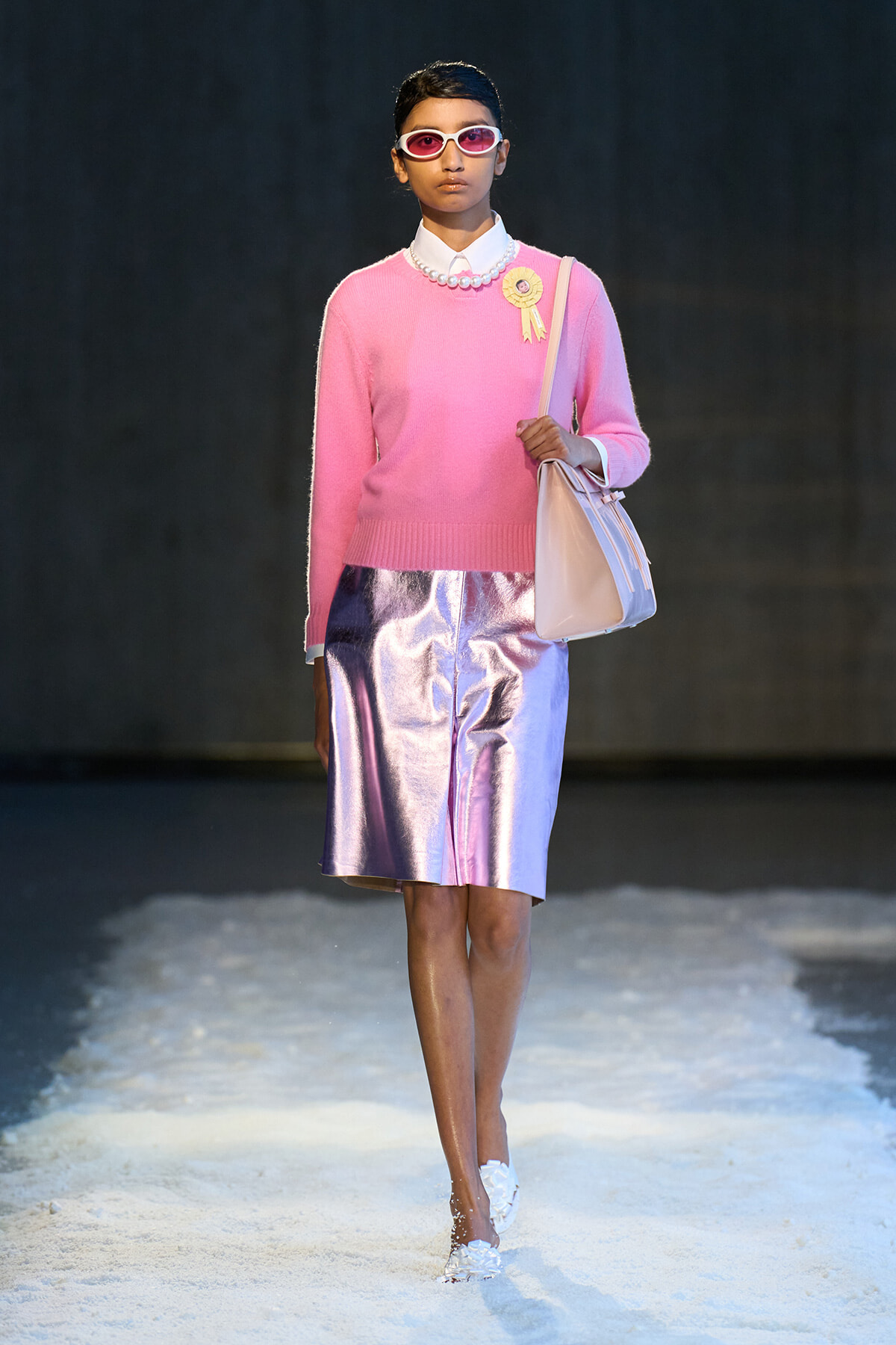 Model walking a runway in a pink knit sweater, pearl necklace, ribbon badge, iridescent pink-metallic skirt, white shoes, and light pink bag with sunglasses.