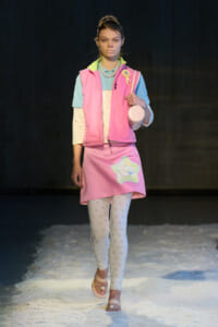 Model walking a fashion runway in pastel pink vest over a light blue top, with a pink star-etched skirt and white patterned tights, holding a small round bag.