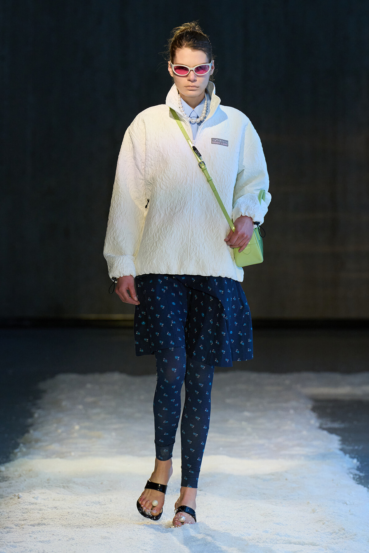 Model walking a fashion runway in a white quilted pullover, navy printed leggings, and a navy skirt, with pink sunglasses and a lime-green crossbody bag.