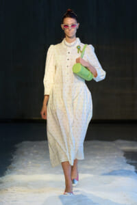 Model walking a runway in a white floral dress with puff sleeves, pink sunglasses, pearl necklace, and a lime-green cylindrical bag close to her chest.