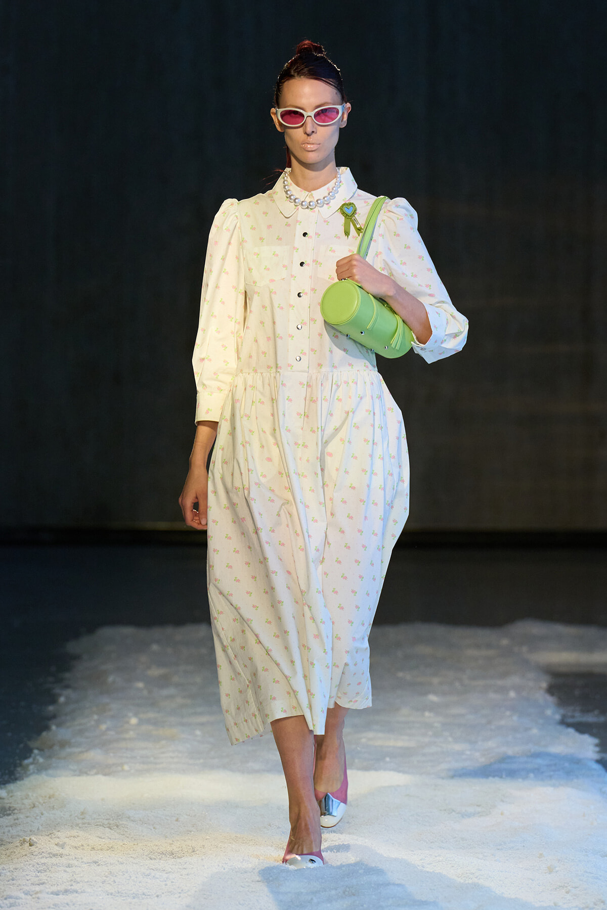 Model walking a runway in a white floral dress with puff sleeves, pink sunglasses, pearl necklace, and a lime-green cylindrical bag close to her chest.