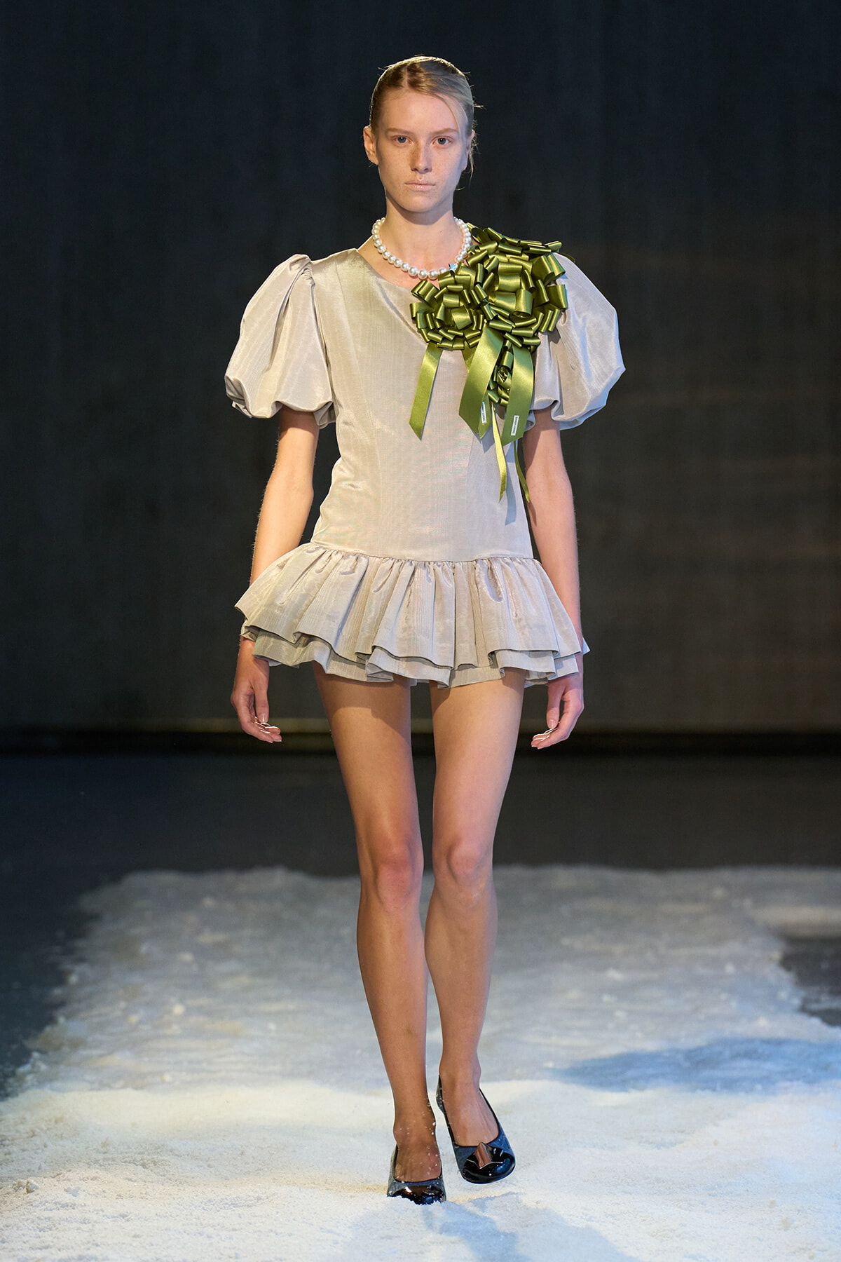 Model walking runway in a pale, puff-sleeve mini dress with a ruffled hem and a large green ribbon bouquet on her shoulder, wearing a pearl necklace.