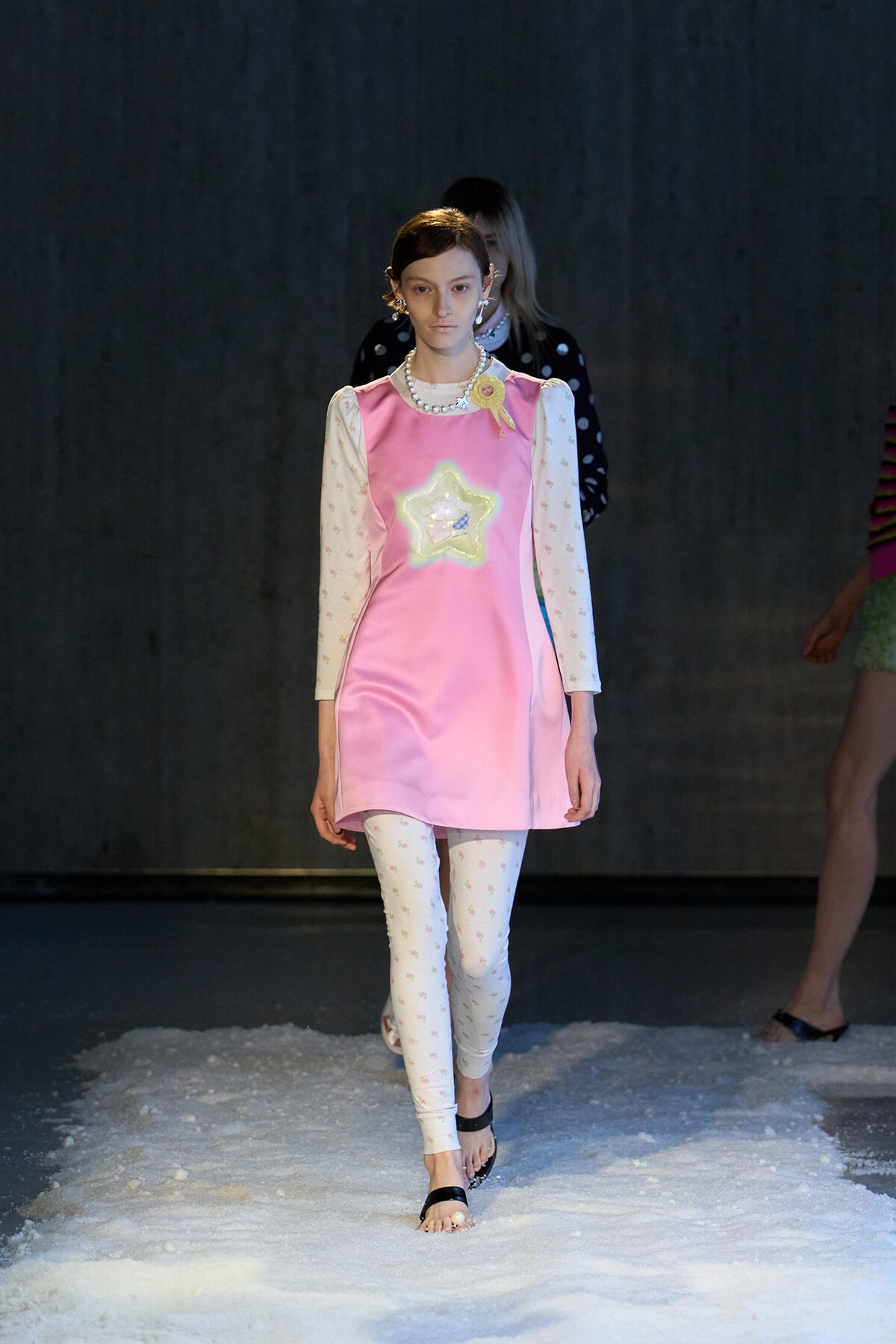 Model walking a snowy runway in a pink satin shift dress with a star appliqué, floral long sleeves, floral leggings, and black sandals on white snow.