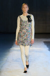 Model on a runway wearing a silver iridescent fringe dress over a pale floral long-sleeve bodysuit, with a black ribbon badge pinned at the shoulder.