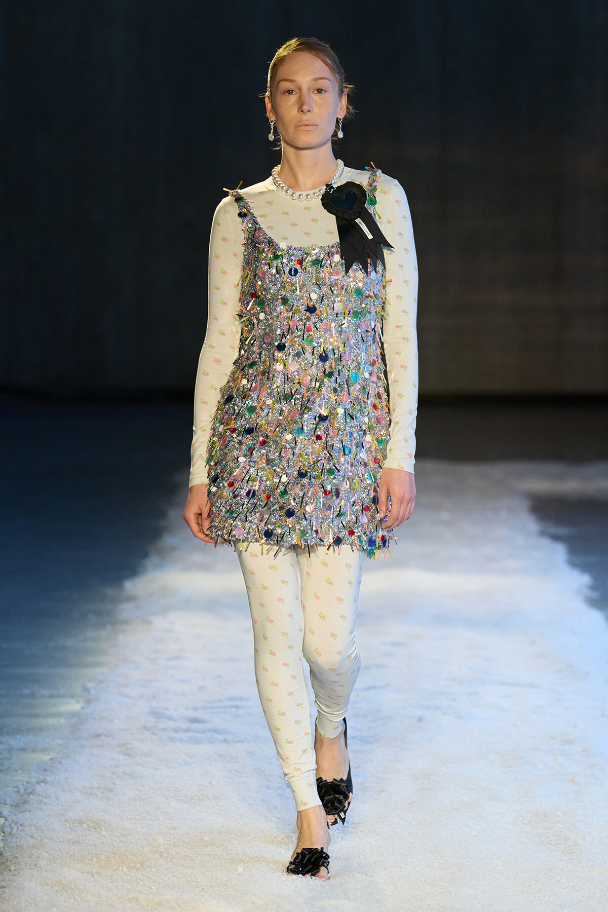 Model on a runway wearing a silver iridescent fringe dress over a pale floral long-sleeve bodysuit, with a black ribbon badge pinned at the shoulder.