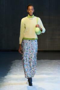 Runway model in a pale yellow sweater, green shirt, pearl necklace, lime bag, and a shimmering, multi-colored fringe skirt.