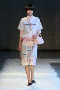 Model on a runway wearing a white graphic tee over a pink striped shirt, a pastel calendar-print skirt, pale pink handbag, and a pearl necklace on a snowy runway.