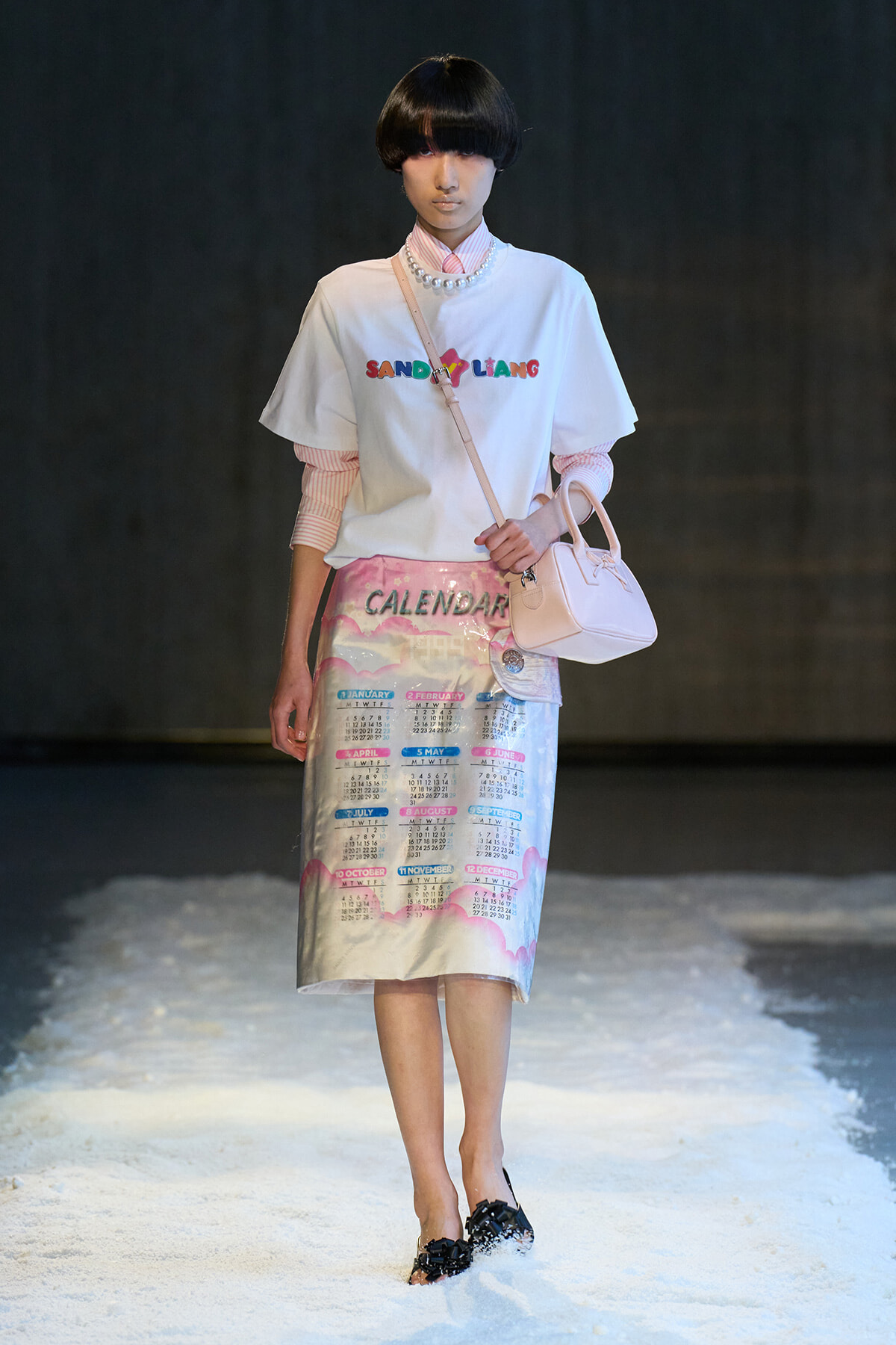 Model on a runway wearing a white graphic tee over a pink striped shirt, a pastel calendar-print skirt, pale pink handbag, and a pearl necklace on a snowy runway.