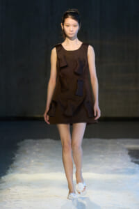 Model walking a fashion show in a sleeveless dark brown A-line dress with sculptural fabric patches and white sandals on a light runway surface