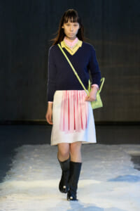 Model walking a fashion show on a snowy runway in a navy sweater, pink shirt, pink gingham pleated skirt, black knee-high boots, and a lime crossbody bag.