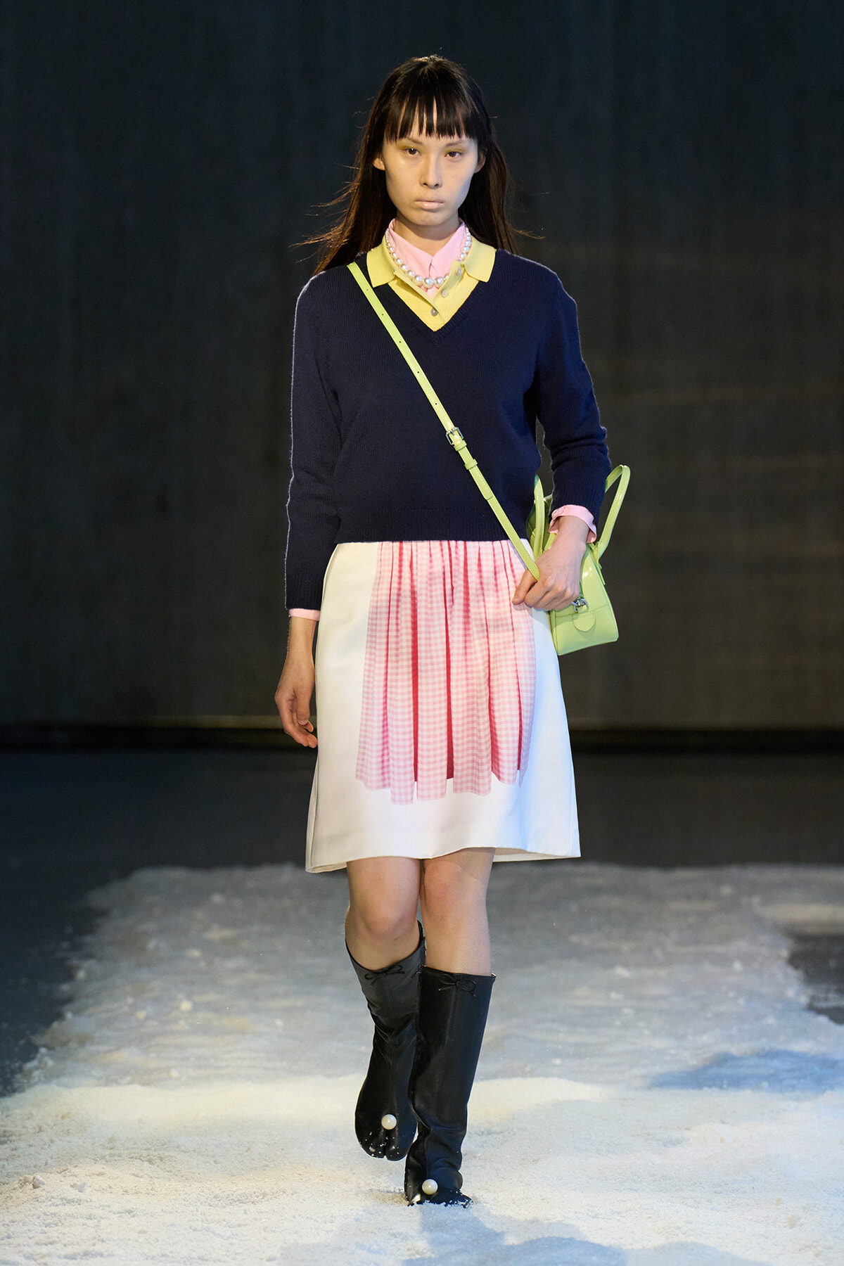 Model walking a fashion show on a snowy runway in a navy sweater, pink shirt, pink gingham pleated skirt, black knee-high boots, and a lime crossbody bag.