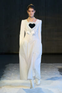 Model in a white floor-length dress with a heart-shaped black chest piece, ruffled edges and ribbon details, on a snowy runway wearing a pearl necklace.