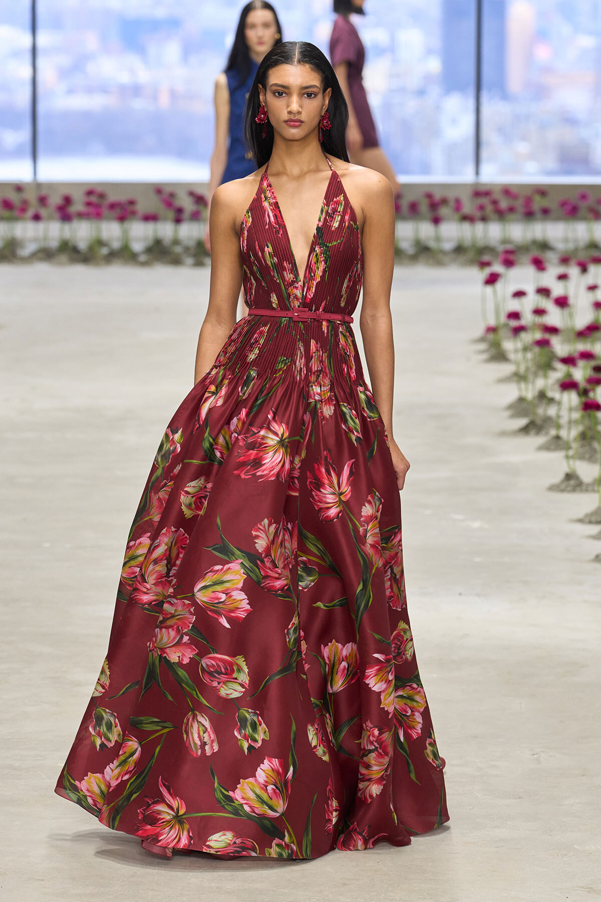 Model wearing a burgundy floral halter gown with a belted waist on a fashion runway.