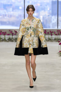 Model walking the runway in a cream floral brocade cape with black velvet hem and matching short dress, black heels.