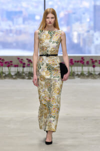 Model walking a runway in a sleeveless floral champagne gown with a black belt and black clutch, urban backdrop behind glass walls.