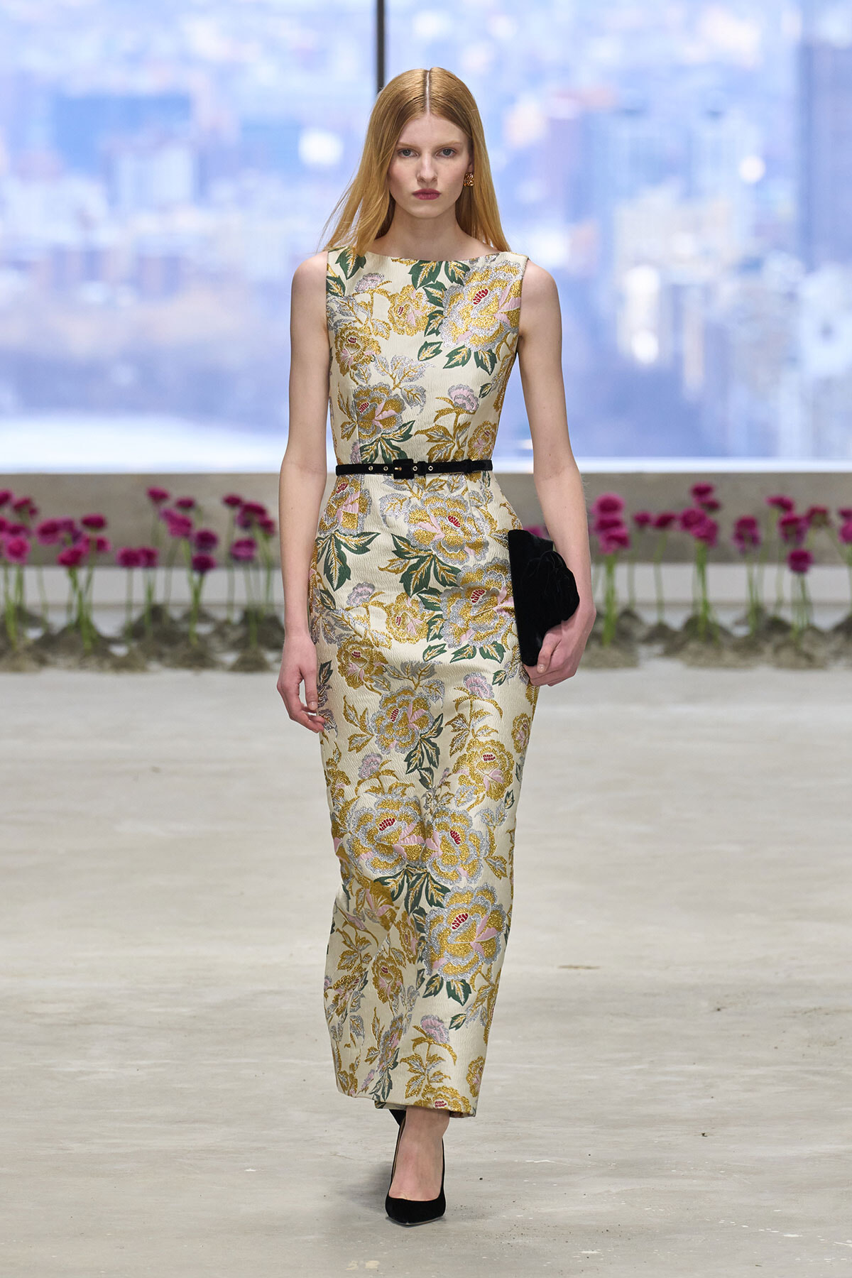 Model walking a runway in a sleeveless floral champagne gown with a black belt and black clutch, urban backdrop behind glass walls.