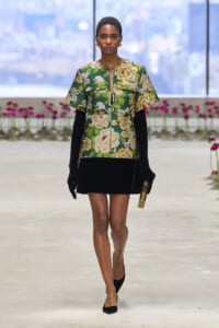 Model walks a fashion runway in a green floral top with gold accents, black mini skirt, black gloves, and a gold clutch.