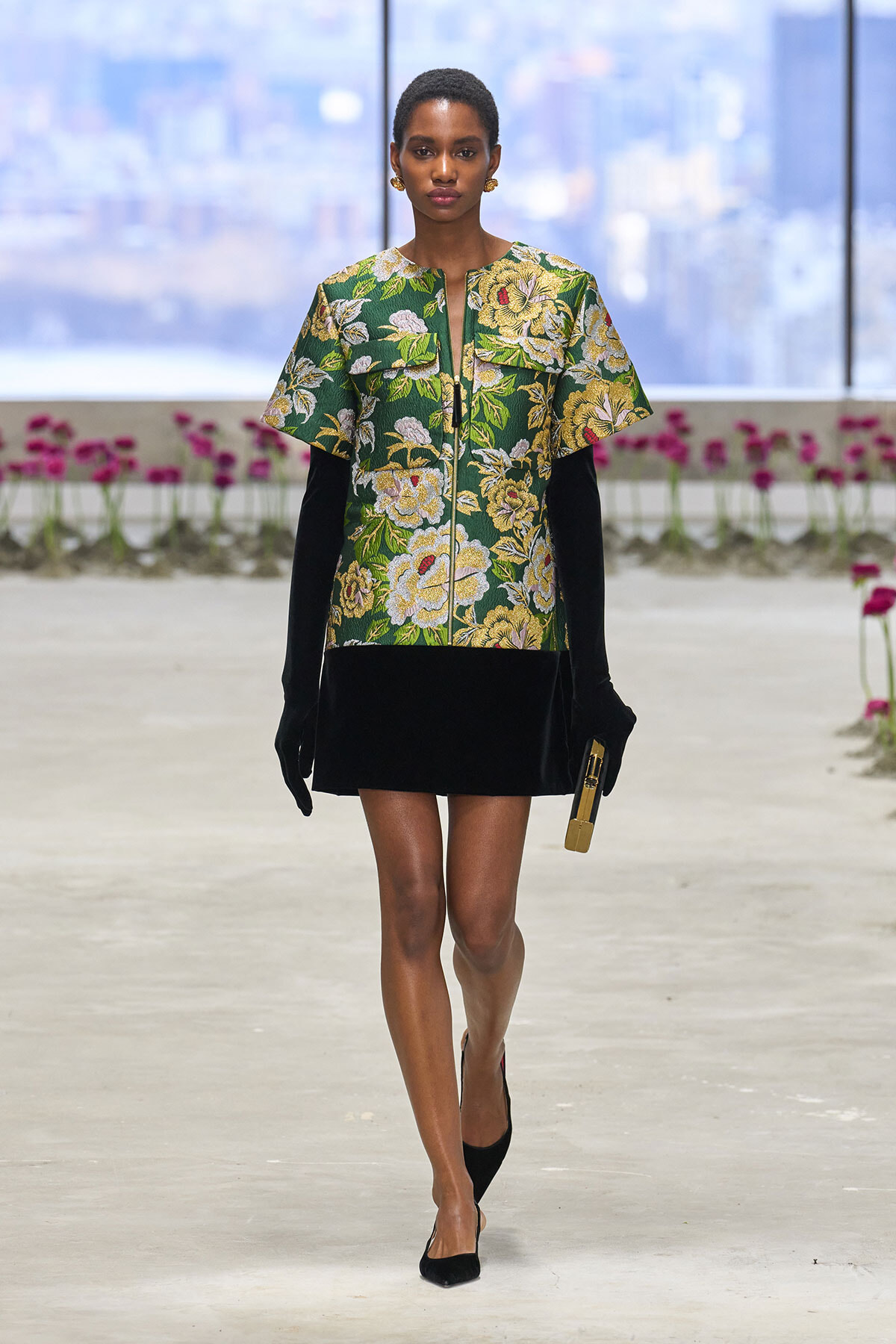 Model walks a fashion runway in a green floral top with gold accents, black mini skirt, black gloves, and a gold clutch.