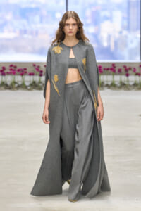 Model walks the runway in a long gray cape with gold floral embroidery over a gray bralette and wide-leg trousers.