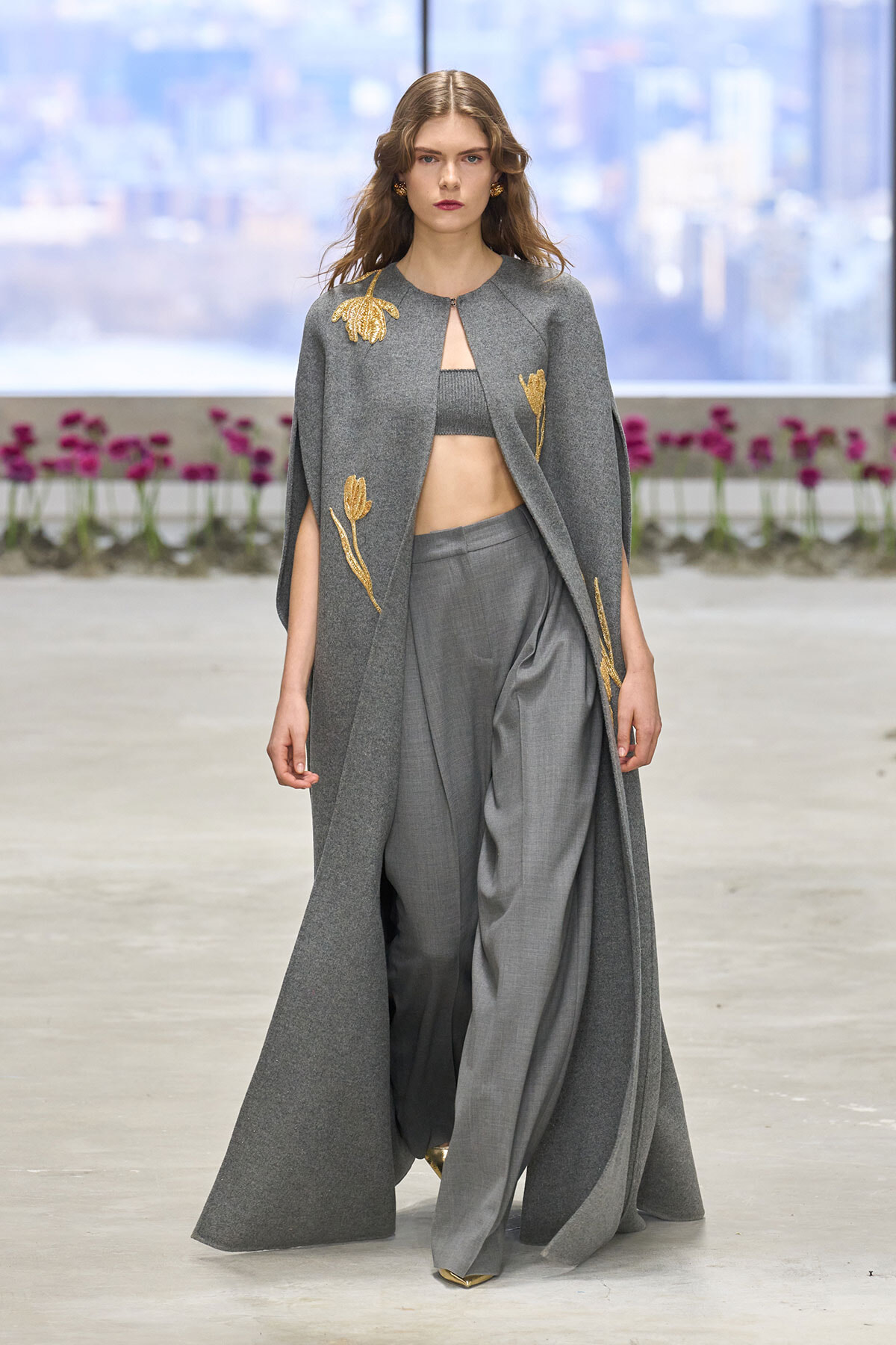 Model walks the runway in a long gray cape with gold floral embroidery over a gray bralette and wide-leg trousers.