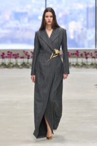 Model wearing a dark gray pinstripe suit with a gold floral brooch at the waist, walking on a runway.